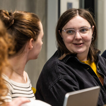 Two people engaged in a work conversation - a woman with her hair up in a bun listening attentively, and a woman in a dark jacket with glasses and a yellow top, mid-sentence and clearly making a point, with a laptop visible in the foreground.