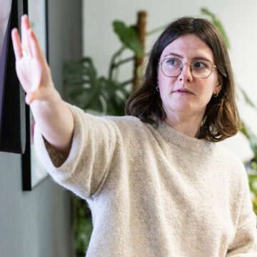 A woman with glasses in a cream jumper, hand raised mid-gesture as though making a decisive point in a presentation or meeting, with a leafy plant visible in the background.