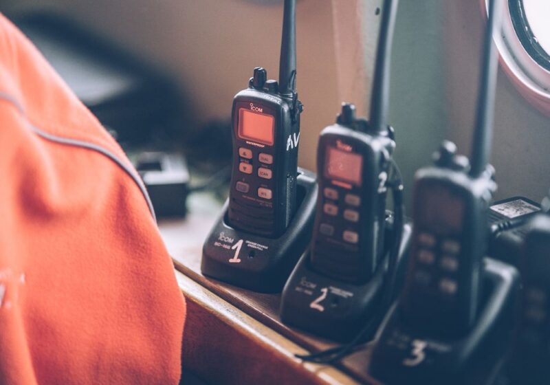 Three handheld Icom radio devices docked in charging stations, with an orange high-visibility jacket visible in the foreground - critical communications equipment of the kind used by emergency services.
